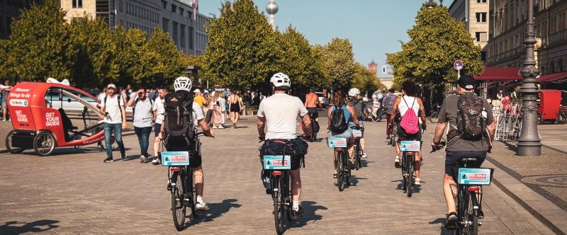 Cyclists riding through an urban area with trees and buildings, a tower visible in the background.