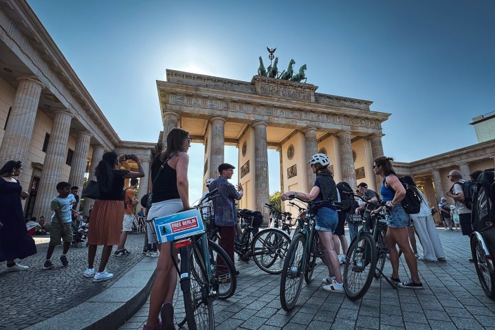 People with bicycles gathered at the Brandenburg Gate in Berlin on a sunny day.