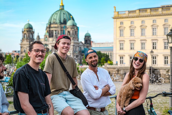 Four people smiling outdoors with a dog, city buildings in background.