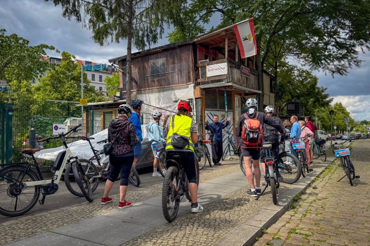 Cyclists gathered in front of a rustic building on a city street, wearing helmets and standing with their bikes.