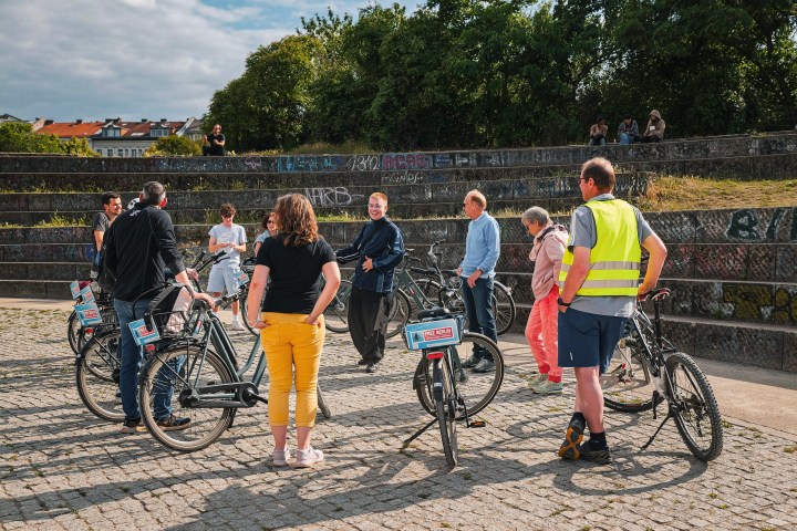 People with bicycles gather in a park by graffiti-covered steps, with trees and buildings in the background.