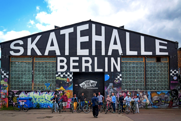 Group of people with bikes outside Skatehalle Berlin building with graffiti.