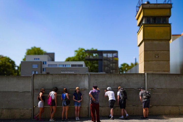 People peer through gaps in a concrete wall at Berlin Wall Memorial on a sunny day.