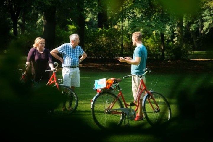 Three people with bicycles talking in a sunny park with trees in the background.