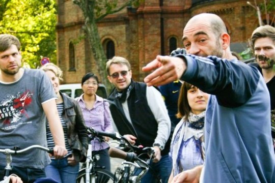 A man gestures while speaking to a group of people with bicycles outside.