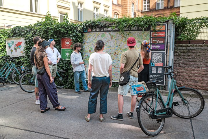 Group of people looking at a map near bicycles and a brick wall with greenery.