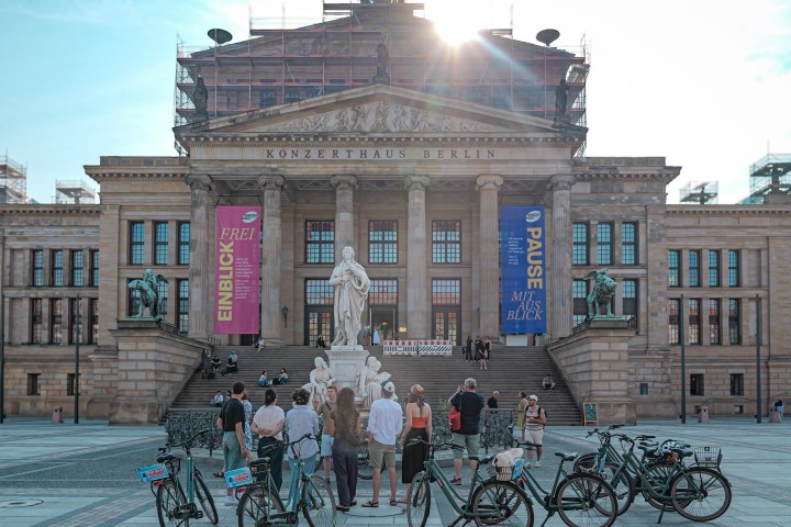 People and bicycles in front of the Konzerthaus Berlin with sunlight behind the building.