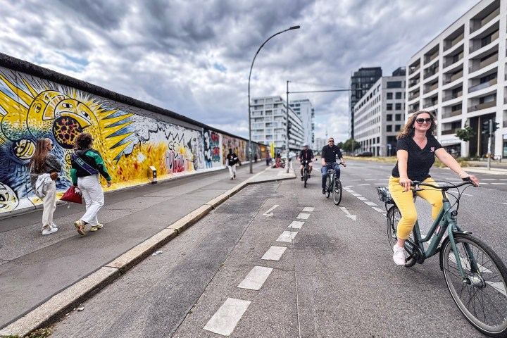 People biking and walking beside a street art-covered wall in an urban area.