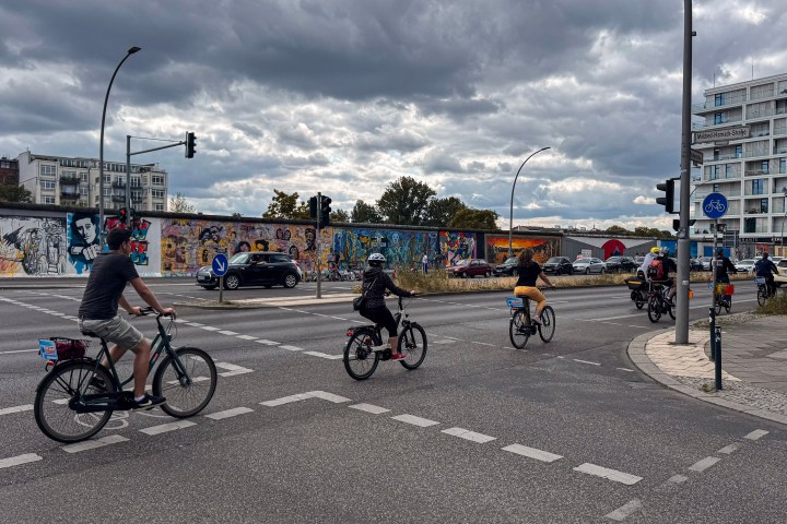 Cyclists riding near a vibrant graffiti-covered wall and a busy intersection under a cloudy sky.