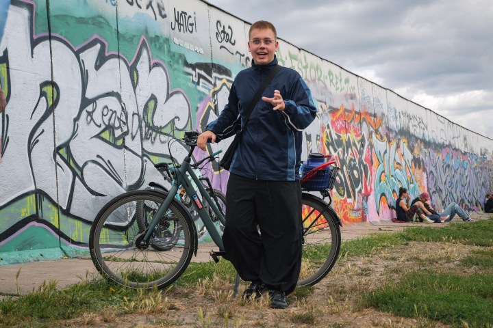 Person with a bike in front of a graffiti-covered wall, sky is cloudy.