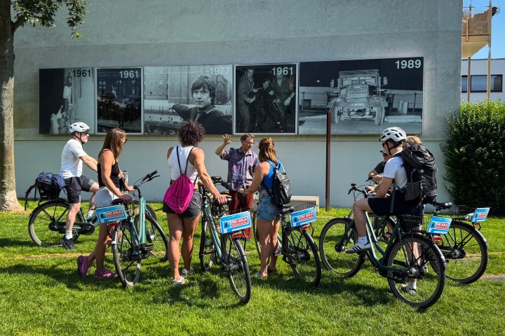 Group of cyclists gathered near outdoor photo exhibit on building wall.