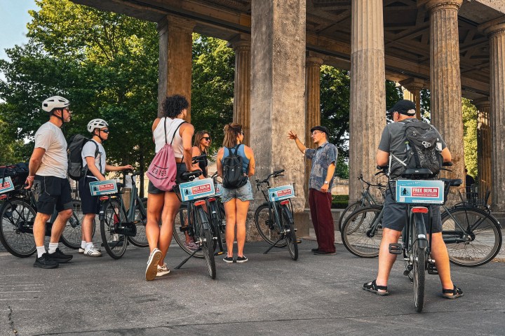 Cyclists gathered around a guide in front of an ancient-style colonnade.