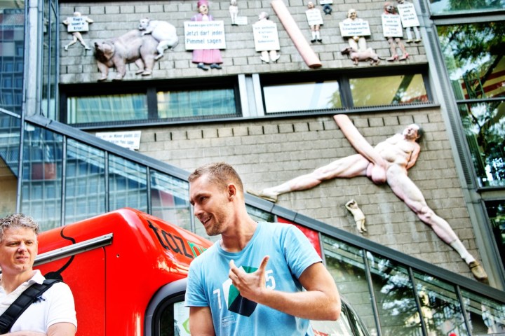 Two men stand near a red vehicle; a wall with quirky sculptures is in the background.