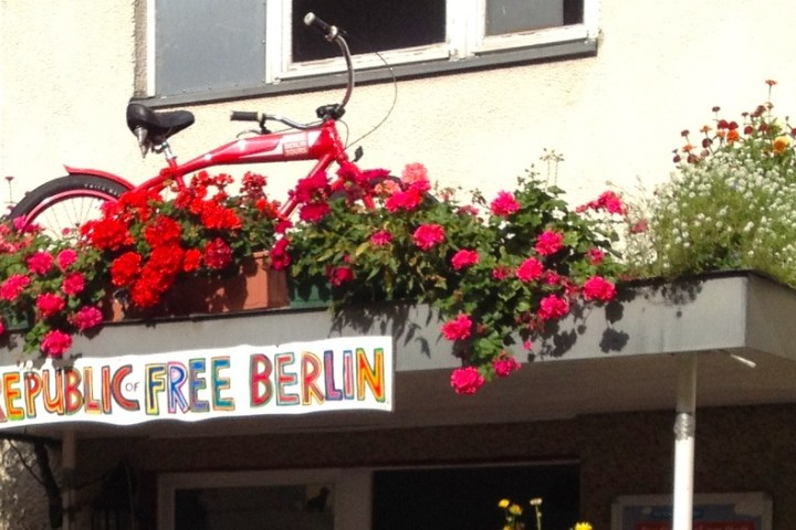 Red bicycle on awning with 'Republic of Free Berlin' sign, surrounded by colorful flowers.
