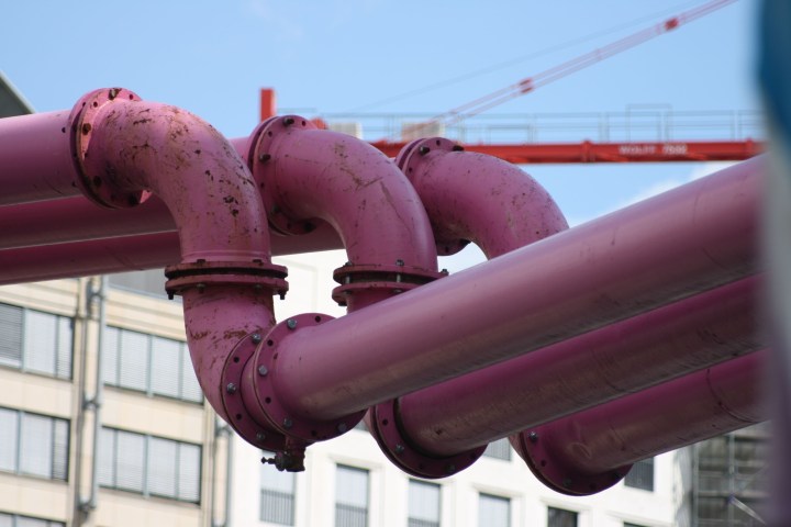 Pink pipes with flanges on a city street, crane and buildings in background.