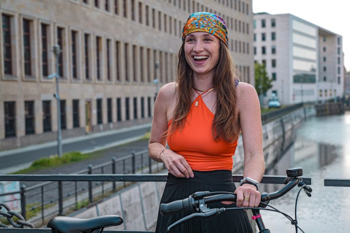 Smiling woman with headband and orange top stands with a bike on a canal bridge.
