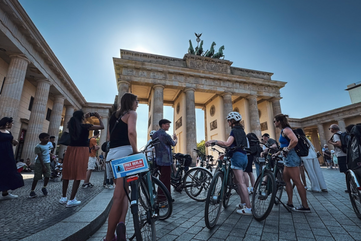 People with bicycles gathered at Brandenburg Gate in sunny weather.