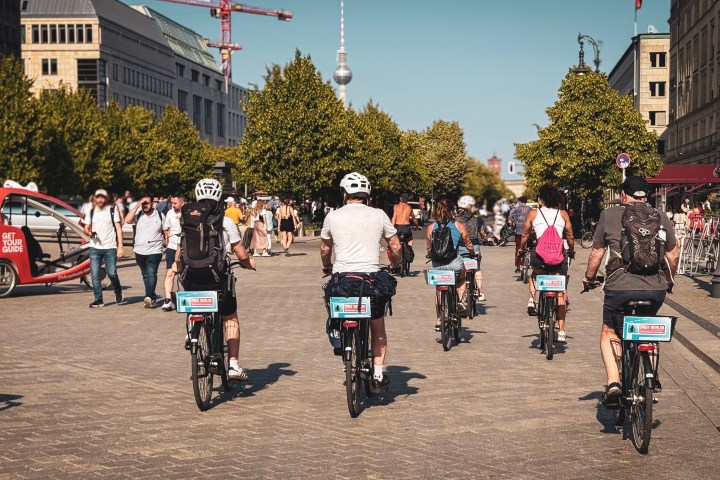 Cyclists riding in a busy city square under a clear sky with trees and buildings nearby.