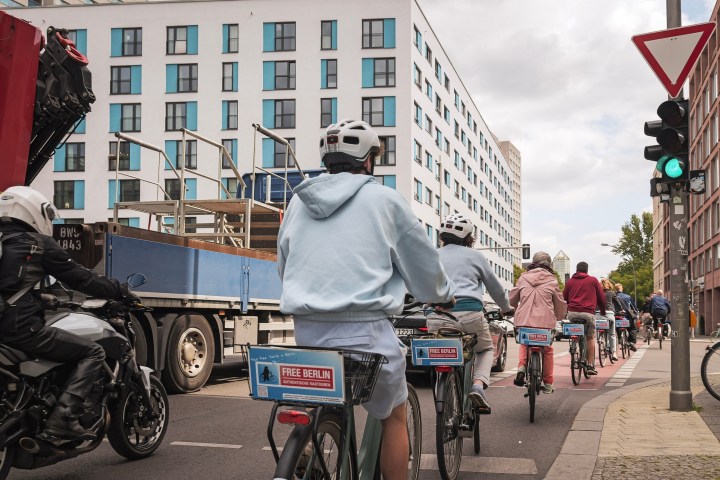 Cyclists in helmets ride in a city lane beside a truck and motorbike.