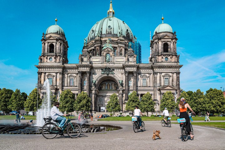 People biking near a large cathedral with a fountain and a small dog in the foreground.