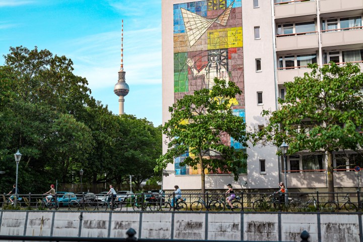 Urban scene with a mural on a building and a TV tower in the background, surrounded by trees and cyclists.