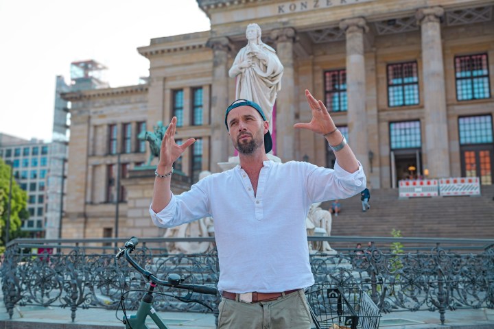 Man in white shirt gestures in front of Konzerthaus Berlin, with a bicycle nearby.
