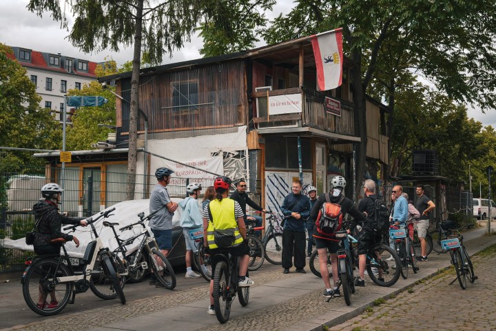 Group of cyclists with helmets gathered by a rustic wooden building and trees.
