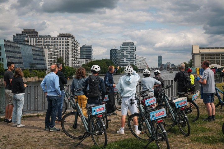Group of cyclists with helmets stand by a river, facing modern buildings on a cloudy day.