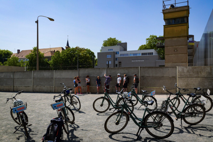 Bicycles parked near a wall with a watchtower and people standing nearby under a clear blue sky.