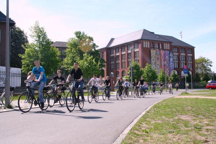 Group of cyclists riding on a sunny day near a large building with banners.