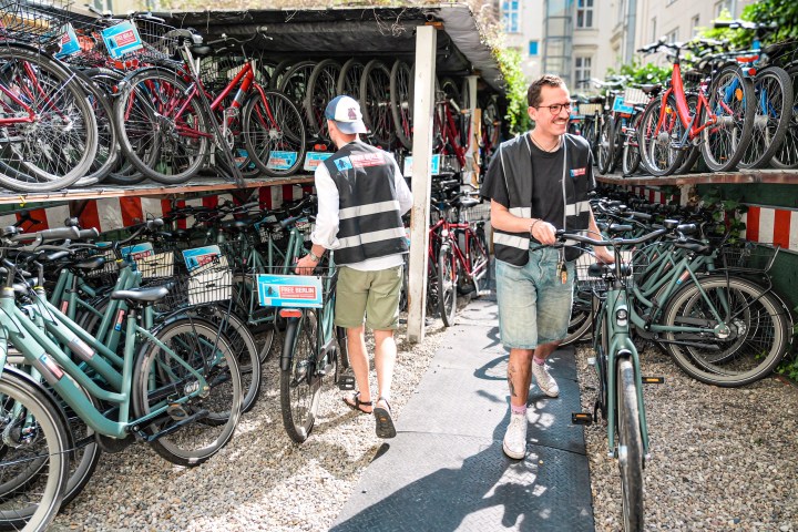 Two people in a bike rental area, walking with bicycles among many parked bikes.