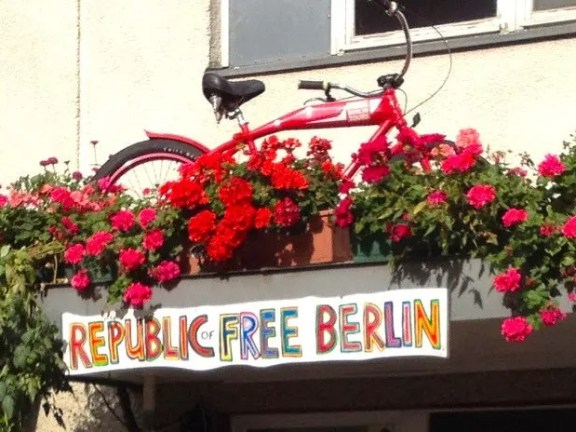 Red bicycle and flowers on balcony above 'Republic of Free Berlin' sign.