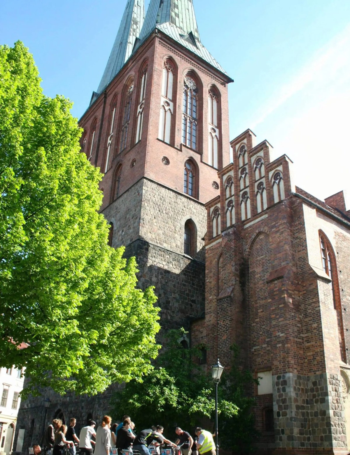 People gathered near a tall brick church tower on a sunny day.
