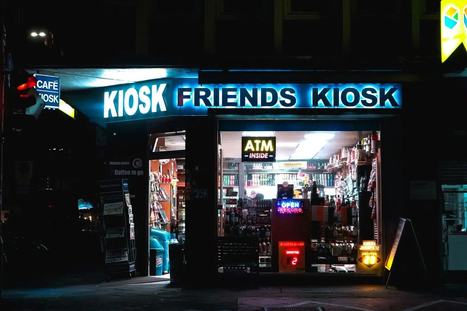 Night view of a brightly lit kiosk with signs for ATM, open, and various products inside.